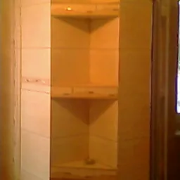 Wnęka narożna z półkami wyłożona jasnymi, kwadratowymi płytkami ceramicznymi w łazience. Widoczne trzy półki z dekoracyjnym obramowaniem. Niska jakość zdjęcia.